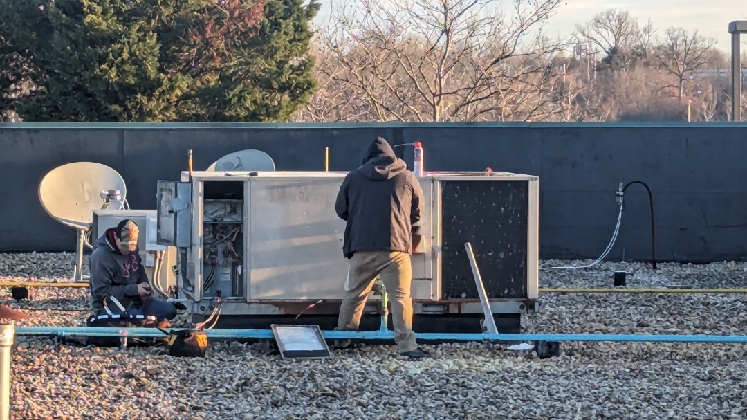 Two HVAC technicians service a rooftop dehumidification system surrounded by satellite dishes and gravel.