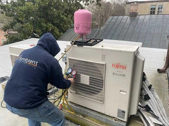 A technician in a hoodie checks gauges on a Fujitsu HVAC unit on a rooftop, with a pink refrigerant tank placed on top of the unit.